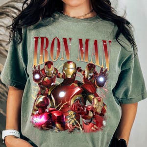 Puede incluir: Camiseta verde oliva con un gr&aacute;fico de Iron Man en varias poses y el texto "IRON MAN". El gr&aacute;fico incluye figuras de Iron Man rojas y doradas con detalles de rayos.