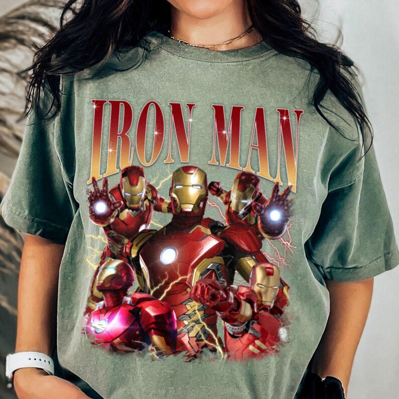 Puede incluir: Camiseta verde oliva con un gr&aacute;fico de Iron Man en varias poses y el texto "IRON MAN". El gr&aacute;fico incluye figuras de Iron Man rojas y doradas con detalles de rayos.