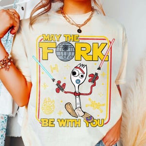 May include: Cream-colored t-shirt with a graphic featuring a fork character holding lightsabers, with the text "MAY THE FORK BE WITH YOU" in yellow and red. The design includes a Death Star and other Star Wars-themed elements.