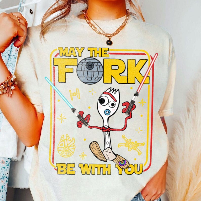May include: Cream-colored t-shirt with a graphic featuring a fork character holding lightsabers, with the text "MAY THE FORK BE WITH YOU" in yellow and red. The design includes a Death Star and other Star Wars-themed elements.