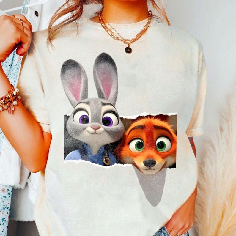 May include: Cream-colored t-shirt featuring a graphic of Judy Hopps and Nick Wilde from Zootopia. Judy, a gray rabbit with purple eyes, and Nick, an orange fox with green eyes, are depicted in a torn paper effect.