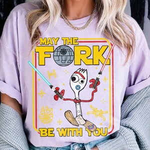 May include: A light purple t-shirt with a graphic featuring a fork character holding lightsabers. The text reads "MAY THE FORK BE WITH YOU" in yellow and white. The design includes a Death Star and other Star Wars elements.