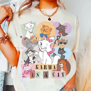 Puede incluir: Una camiseta beige claro con un estampado gr&aacute;fico de varios gatos de dibujos animados. El gr&aacute;fico presenta un gato blanco con orejas rosas y un lazo rosa, un gato negro con un collar rojo y un gato marr&oacute;n con un collar amarillo. El texto "KARMA IS A CAT" est&aacute; impreso en letras negras en la camiseta.