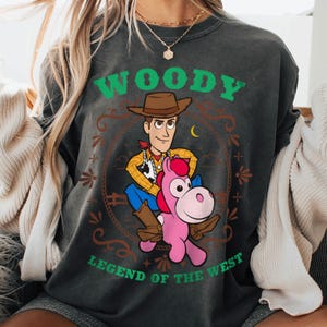 May include: A charcoal gray crewneck t-shirt featuring a graphic of Woody from Toy Story riding a pink horse. The text reads WOODY at the top and LEGEND OF THE WEST at the bottom in green letters. The design includes a brown cowboy hat and lasso.