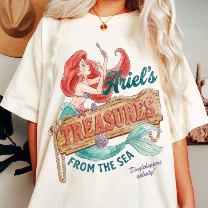 May include: Cream-colored t-shirt featuring a watercolor illustration of Ariel from The Little Mermaid. The design includes Ariel, a wooden sign that reads "Ariel's Treasures From The Sea," and the phrase "Dinglehoppers aplenty!"