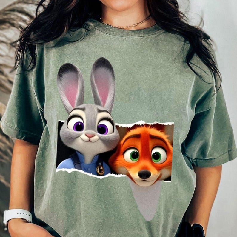 May include: Sage green t-shirt featuring a graphic of Judy Hopps and Nick Wilde from Zootopia. Judy, a gray rabbit, is in a blue uniform. Nick, an orange fox, has green eyes. The graphic is designed to look like it's peeking through a torn paper.