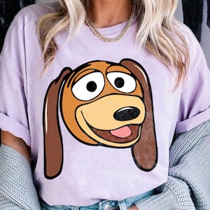 Pu&ograve; includere: Una maglietta lilla con un'immagine a cartoni animati di Slinky Dog di Toy Story. La grafica mostra la faccia di Slinky con pelo marrone e marrone chiaro, grandi occhi bianchi e una lingua rosa. La maglietta ha maniche corte.