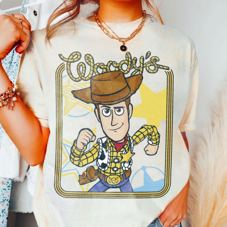 May include: A cream-colored short-sleeve t-shirt featuring a graphic print of the character Woody from Toy Story. The design includes the text Woody's in a rope-style font, a yellow star background, and a brown cowboy hat, yellow shirt, and vest.