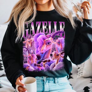 May include: Black sweatshirt featuring a purple graphic of a cartoon gazelle with purple hair and the word "GAZELLE" in a purple font. The gazelle is holding a microphone. The sweatshirt is made of a soft material.