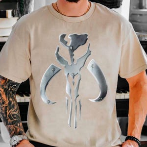 Puede incluir: Camiseta beige con un emblema plateado met&aacute;lico. El emblema es un dise&ntilde;o sim&eacute;trico con elementos curvos y un componente vertical central. La camiseta tiene mangas cortas y cuello redondo.
