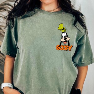 Puede incluir: Camiseta verde salvia con un dibujo animado de Goofy y la palabra "GOOFY" en naranja. El dise&ntilde;o est&aacute; en la parte superior izquierda del pecho. La camiseta tiene mangas cortas y un corte holgado.