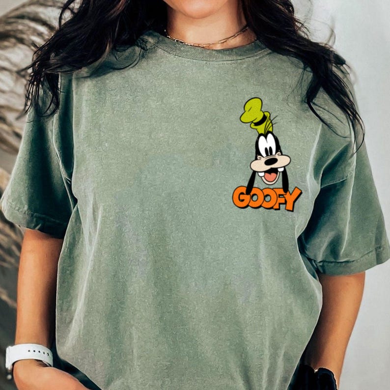 Puede incluir: Camiseta verde salvia con un dibujo animado de Goofy y la palabra "GOOFY" en naranja. El dise&ntilde;o est&aacute; en la parte superior izquierda del pecho. La camiseta tiene mangas cortas y un corte holgado.