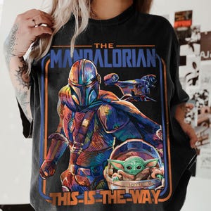 May include: Black t-shirt featuring a colorful graphic of The Mandalorian, a character from the Star Wars series, with the text "THE MANDALORIAN" and "THIS IS THE WAY". The design includes the Mandalorian, a ship, and Grogu.