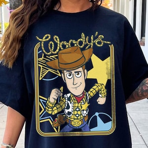 May include: A black short-sleeved t-shirt featuring a graphic print of a cowboy character wearing a brown hat, a yellow plaid shirt, and a sheriff badge. The text "Woody's" appears at the top in a rope-style font, framed by a yellow border.