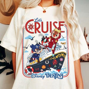 K&ouml;nnte beinhalten: Ein cremefarbenes T-Shirt mit einer Grafik eines Kreuzfahrtschiffs und zwei Streifenh&ouml;rnchen als Superhelden. Der Text lautet Let's CRUISE und Disney DESTINY. Das Schiff ist mit verschiedenen Superhelden-Emblemen und einem Rettungsring verziert.