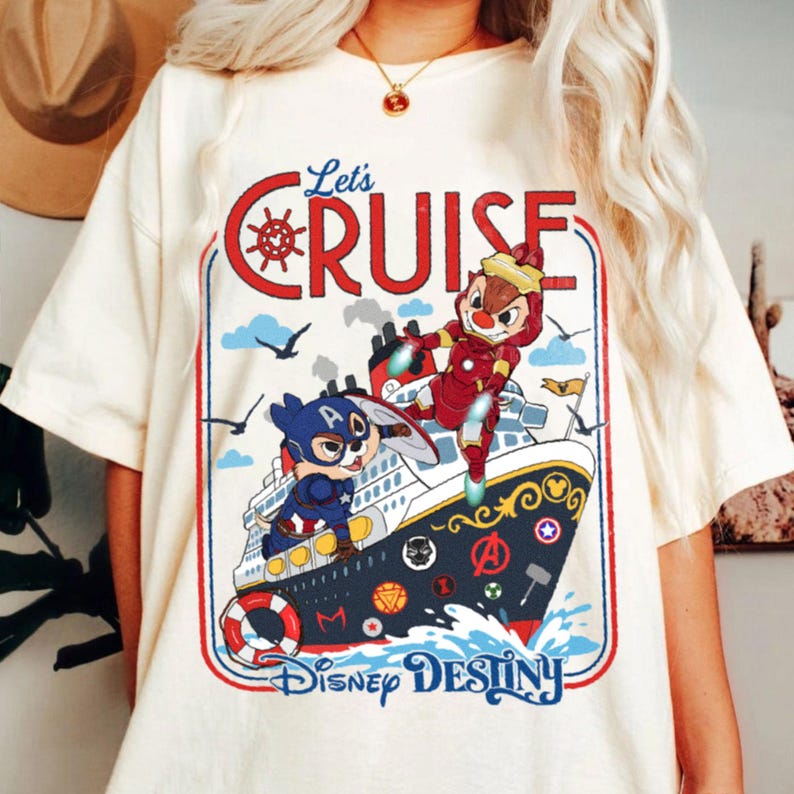 K&ouml;nnte beinhalten: Ein cremefarbenes T-Shirt mit einer Grafik eines Kreuzfahrtschiffs und zwei Streifenh&ouml;rnchen als Superhelden. Der Text lautet Let's CRUISE und Disney DESTINY. Das Schiff ist mit verschiedenen Superhelden-Emblemen und einem Rettungsring verziert.