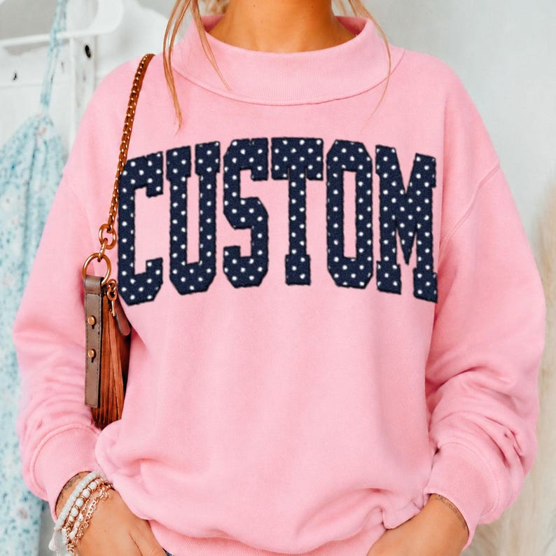 May include: A light pink long-sleeve sweatshirt featuring a mock neck collar. The word CUSTOM is printed across the chest in large, dark navy blue block letters with a white polka dot pattern. The fabric appears to be a soft, solid-colored cotton blend.