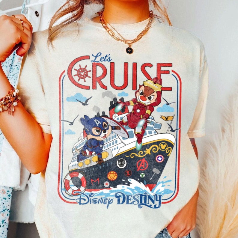 K&ouml;nnte beinhalten: Ein cremefarbenes T-Shirt mit einem Grafikdruck eines Kreuzfahrtschiffes und Charakteren in Superheldenkost&uuml;men. Der Text lautet Let's CRUISE und Disney DESTINY. Das Schiff ist mit verschiedenen Superhelden-Symbolen verziert und rot umrandet.