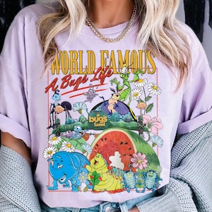 May include: A light purple t-shirt featuring a colorful graphic from the animated film "A Bug's Life." The design includes the text "World Famous A Bug's Life" and various cartoon insects, flowers, and a watermelon.