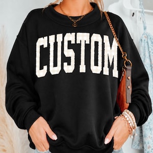 May include: A black long-sleeve sweatshirt featuring the word CUSTOM in large, white, block-style letters with a subtle light-colored checkered pattern. The garment has a ribbed crew neckline and is worn with a gold chain necklace and a gold chain strap bag.