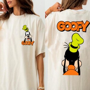 Puede incluir: Camiseta color crema con una imagen de dibujos animados de Goofy en la parte delantera y trasera. La parte delantera muestra la cara de Goofy y la palabra "GOOFY" en naranja. La parte trasera muestra la cabeza de Goofy y la palabra "GOOFY" en naranja.