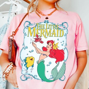 Puede incluir: Camiseta rosa claro con un gr&aacute;fico de Ariel de La Sirenita. El dise&ntilde;o incluye a Ariel con cabello rojo, una cola verde y una flor amarilla, junto con Flounder y Sebasti&aacute;n. Las palabras "The Little Mermaid" son amarillas.