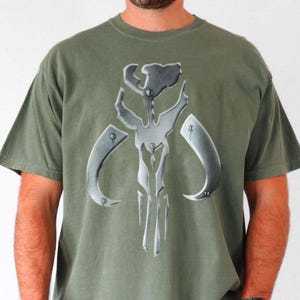 Puede incluir: Camiseta verde oliva con un emblema plateado met&aacute;lico. El emblema es un dise&ntilde;o sim&eacute;trico con elementos curvos y una l&iacute;nea vertical central. La camiseta tiene mangas cortas y cuello redondo.