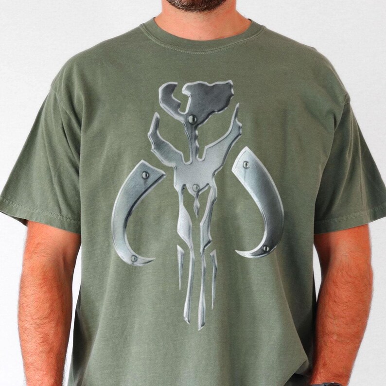 Puede incluir: Camiseta verde oliva con un emblema plateado met&aacute;lico. El emblema es un dise&ntilde;o sim&eacute;trico con elementos curvos y una l&iacute;nea vertical central. La camiseta tiene mangas cortas y cuello redondo.