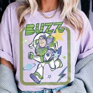 May include: A light purple t-shirt featuring a graphic of the character Buzz Lightyear in a white, green, and purple space suit. The design includes the word BUZZ in green letters, a yellow star, a blue planet, and a blue lightning bolt icon.