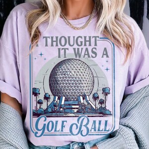 May include: A light purple t-shirt with the text "I THOUGHT IT WAS A GOLF BALL" in a blue and white graphic. The graphic features a large geodesic sphere, trees, and a blue border.