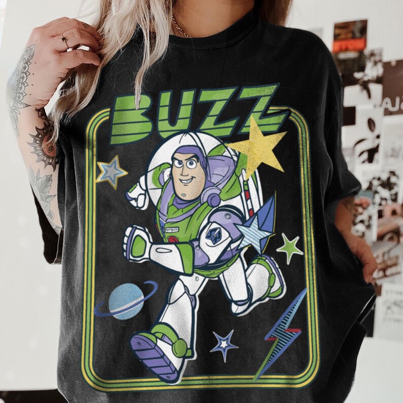 May include: A black short-sleeve t-shirt featuring a graphic print of the character Buzz Lightyear in a space suit. The design includes the text BUZZ in green, surrounded by yellow stars, a blue planet with rings, and a blue lightning bolt icon.
