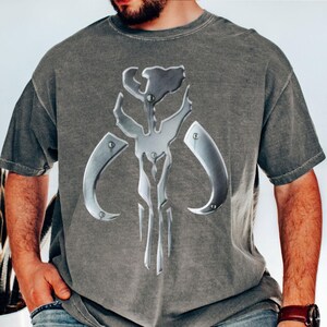 Puede incluir: Camiseta gris con un emblema plateado met&aacute;lico. El emblema es una calavera estilizada con cuernos curvos y detalles angulares. La camiseta tiene mangas cortas y cuello redondo.