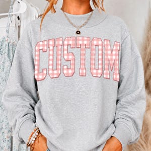 May include: A heather gray long-sleeve sweatshirt featuring the word CUSTOM in large, block-style letters with a pink and white gingham check pattern. The sweatshirt has a crew neckline and is paired with a gold-tone chain necklace with a small pendant.