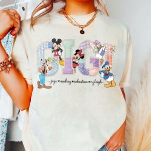 May include: Cream-coloured t-shirt with the word "GIGI" in a patchwork design, featuring Disney characters Mickey Mouse, Minnie Mouse, Goofy, Daisy Duck, and Donald Duck. Names are printed below the characters.