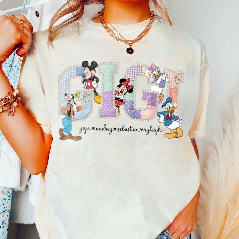 May include: Cream-coloured t-shirt with the word "GIGI" in a patchwork design, featuring Disney characters Mickey Mouse, Minnie Mouse, Goofy, Daisy Duck, and Donald Duck. Names are printed below the characters.