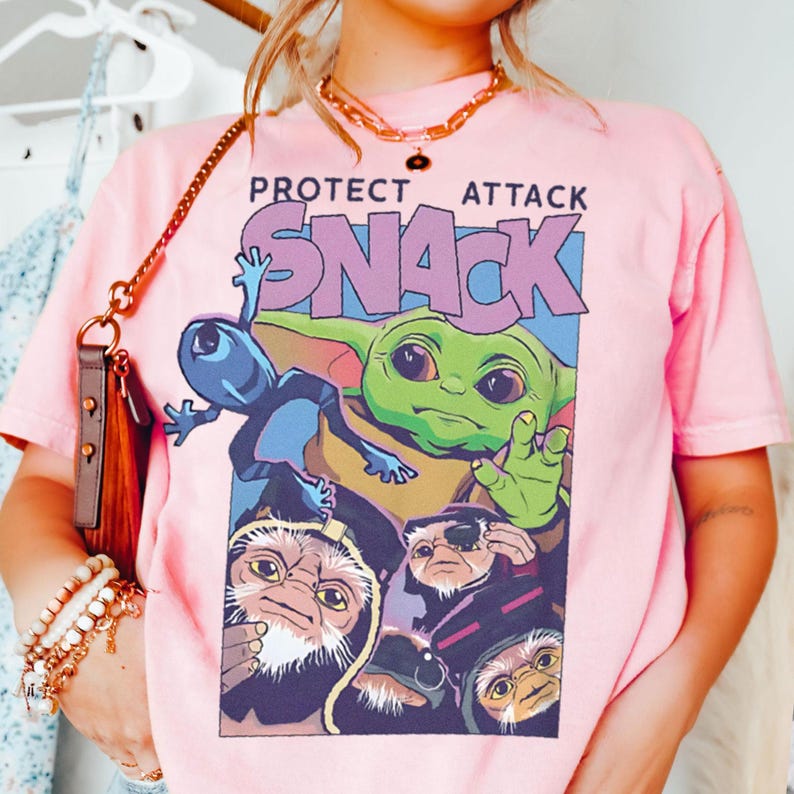 K&ouml;nnte beinhalten: Hellrosa T-Shirt mit einem Aufdruck von Grogu, einem blauen Alien und anderen Charakteren mit den Worten "PROTECT ATTACK SNACK". Das T-Shirt hat kurze &Auml;rmel und einen Rundhalsausschnitt.