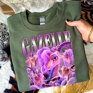May include: Olive green sweatshirt featuring a graphic of cartoon gazelles with purple hair and horns, holding microphones. The word "GAZELLE" is printed in a purple gradient font across the top of the design.
