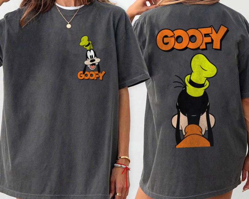 Puede incluir: Camiseta gris oscuro con una imagen de dibujos animados de Goofy. La parte delantera de la camiseta muestra la cara de Goofy con la palabra "GOOFY" en naranja. La parte trasera de la camiseta muestra la cabeza de Goofy y la palabra "GOOFY" en naranja.