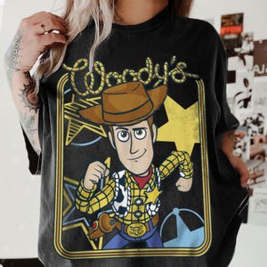 May include: A black t-shirt featuring a graphic print of the character Woody from Toy Story. The design includes the text Woody's in a rope-style font, a yellow star, and a brown cowboy hat. The character wears a yellow plaid shirt.