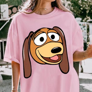 Pu&ograve; includere: T-shirt rosa chiaro con un'immagine a cartoni animati di Slinky Dog di Toy Story. La grafica mostra il volto di Slinky con orecchie marroni, un muso giallo e un'espressione sorridente. La maglietta ha maniche corte e una vestibilit&agrave; ampia.