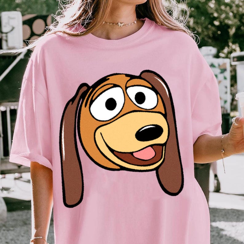 Pu&ograve; includere: T-shirt rosa chiaro con un'immagine a cartoni animati di Slinky Dog di Toy Story. La grafica mostra il volto di Slinky con orecchie marroni, un muso giallo e un'espressione sorridente. La maglietta ha maniche corte e una vestibilit&agrave; ampia.