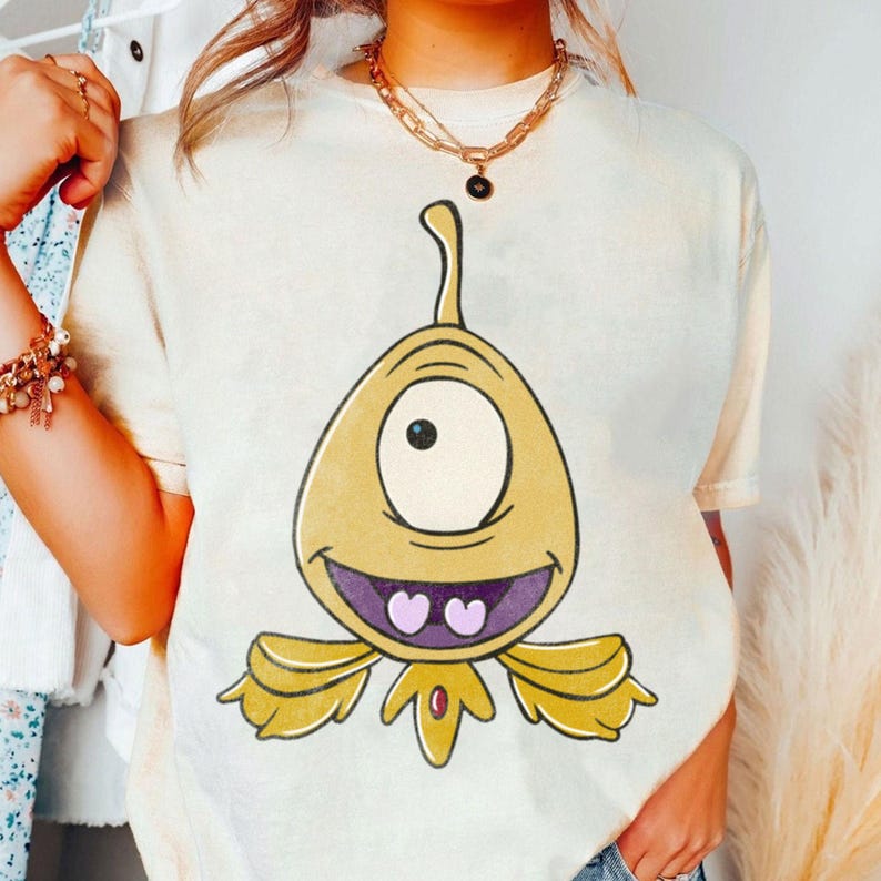 May include: Cream-colored t-shirt featuring a cartoon alien with one eye, a purple mouth, and yellow accents. The alien has a smiling expression and is centered on the shirt. The shirt is a casual, everyday wear item.