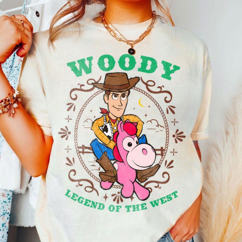 May include: A cream-colored t-shirt featuring a graphic of the character Woody riding a pink horse. The text reads WOODY at the top and LEGEND OF THE WEST at the bottom in green letters. The design is enclosed in a rope-style circular border.