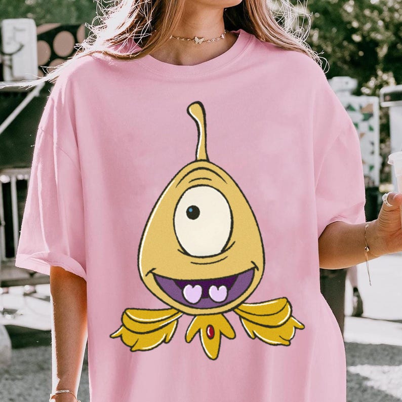 May include: A light pink t-shirt featuring a cartoon alien character. The alien is yellow with a single eye, a smiling mouth, and two heart-shaped teeth. The design includes a curved antenna and wing-like appendages.