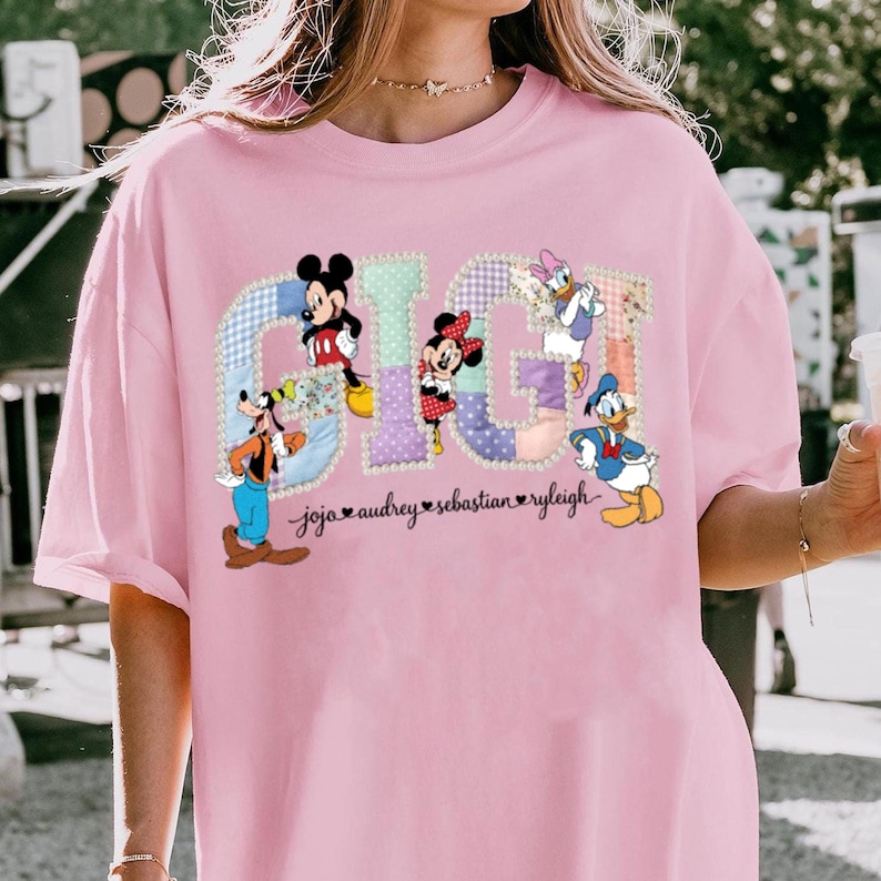 May include: Pale pink t-shirt with the word "GIG" in a patchwork design, featuring Disney characters Mickey Mouse, Minnie Mouse, Donald Duck, Daisy Duck, and Goofy. Names are printed below the word.