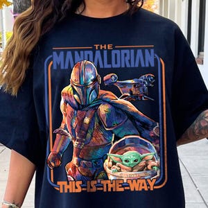 May include: Dark navy blue t-shirt featuring a colorful graphic of The Mandalorian, a spaceship, and Grogu. The text "THE MANDALORIAN" and "THIS IS THE WAY" are displayed in orange and white.