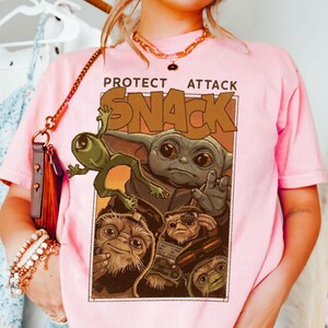 May include: Pink t-shirt featuring a graphic of Baby Yoda and other creatures with the words "PROTECT ATTACK SNACK" in a brown font. The graphic is in a rectangular frame. The shirt has short sleeves.