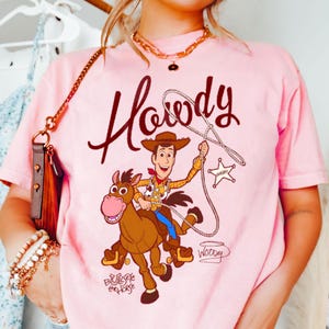 May include: Pink t-shirt featuring a cartoon graphic of Woody from Toy Story riding Bullseye, with the word "Howdy" in a brown script. The design includes a sheriff's badge and the names "Woody" and "Bullseye the Horse".