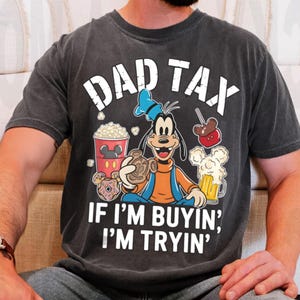 Op de afbeelding: Donkergrijs t-shirt met Goofy-afbeelding en de tekst "DAD TAX" en "IF I'M BUYIN', I'M TRYIN'". Het ontwerp bevat popcorn, een karamelappel, bier en een donut.