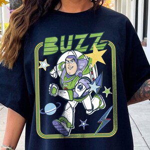 May include: A black short-sleeve t-shirt featuring a graphic of the character Buzz Lightyear in a white, green, and purple space suit. The design includes the word BUZZ in green letters, yellow and blue stars, a planet, and a lightning bolt.